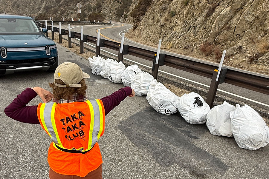 Valerie Botta with orange "Takataka club" vest on, pointing to a row of trash bags